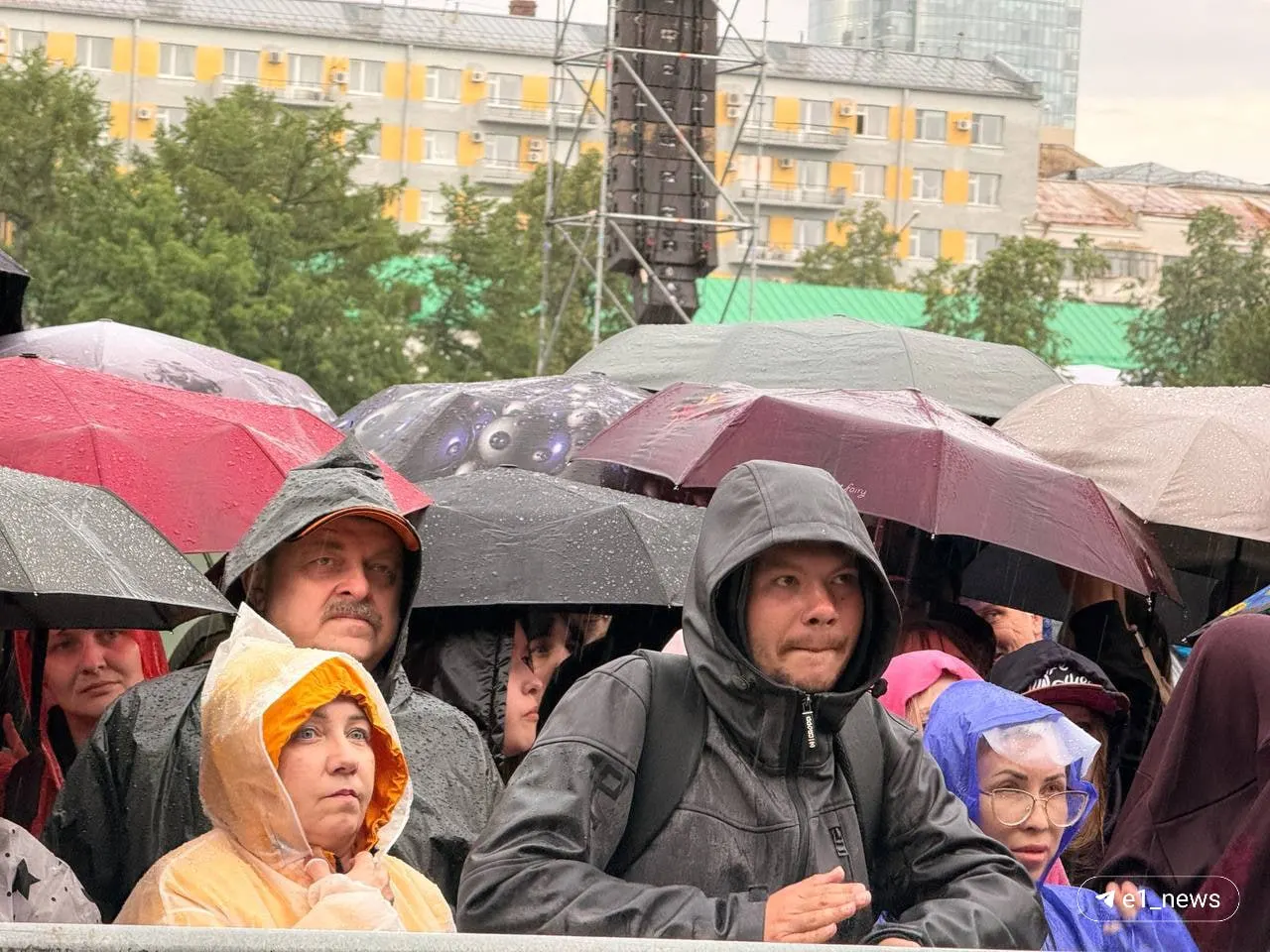 На открытом мероприятии в Екатеринбурге зрители прятались от ливня под зонтиками и дождевиками; выступление Сергея Бобунца задерживается, причина неизвестна.